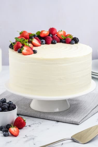 A berry chantilly cake on a cake stand, topped with fresh berries and mint sprigs.