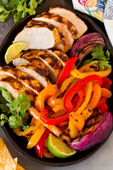 A platter of grilled chicken fajitas with onions, bell peppers, lime wedges and fresh cilantro.