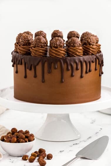 A Nutella cake on a stand with chocolate frosting, a ganache drizzle and chocolate hazelnut candy.