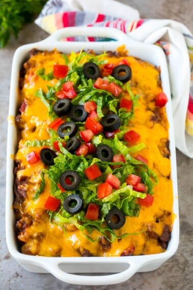 Taco casserole in a baking dish topped with melted cheese, lettuce, tomato and olives.