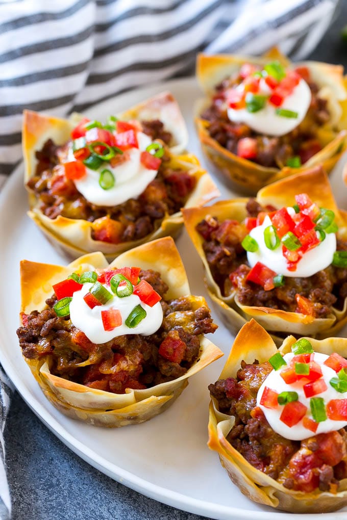 A plate of taco cups made with wontons, seasoned ground beef and cheese, then topped with sour cream, tomatoes and green onions.