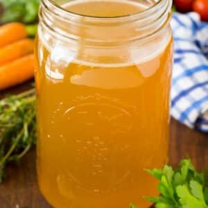 A jar of homemade turkey broth surrounded by herbs and vegetables.