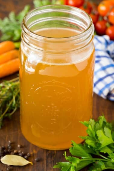 A jar of homemade turkey broth surrounded by herbs and vegetables.